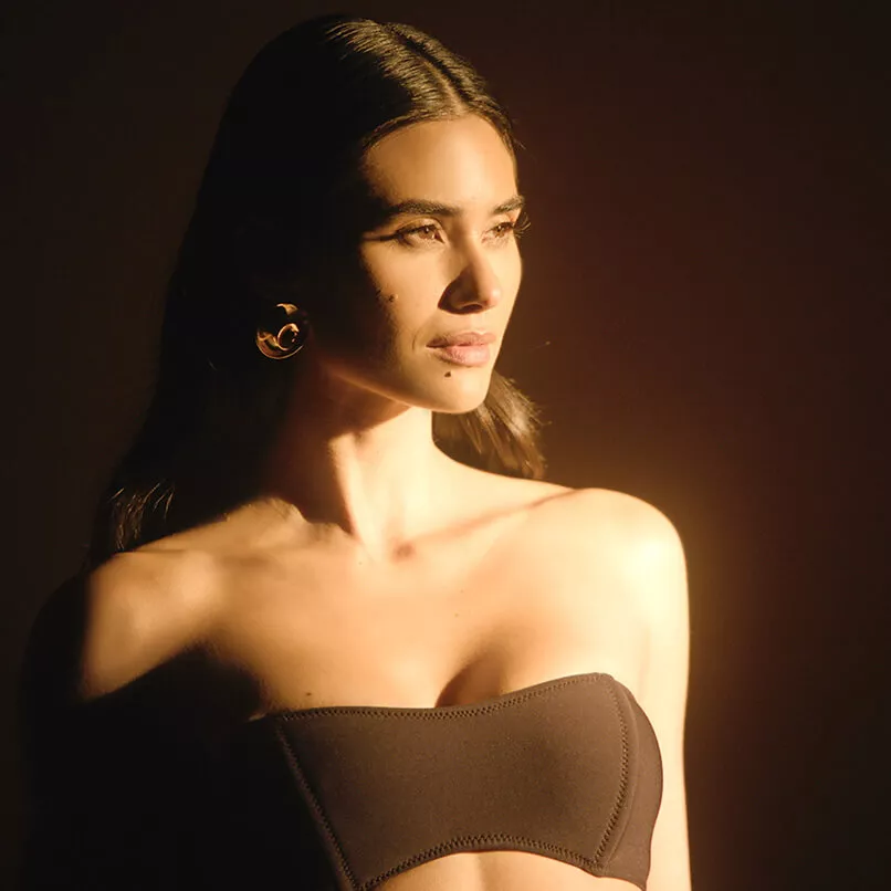 A person with long brunette hair poses in warm afternoon sunlight against a brown background, showing a confident expression.